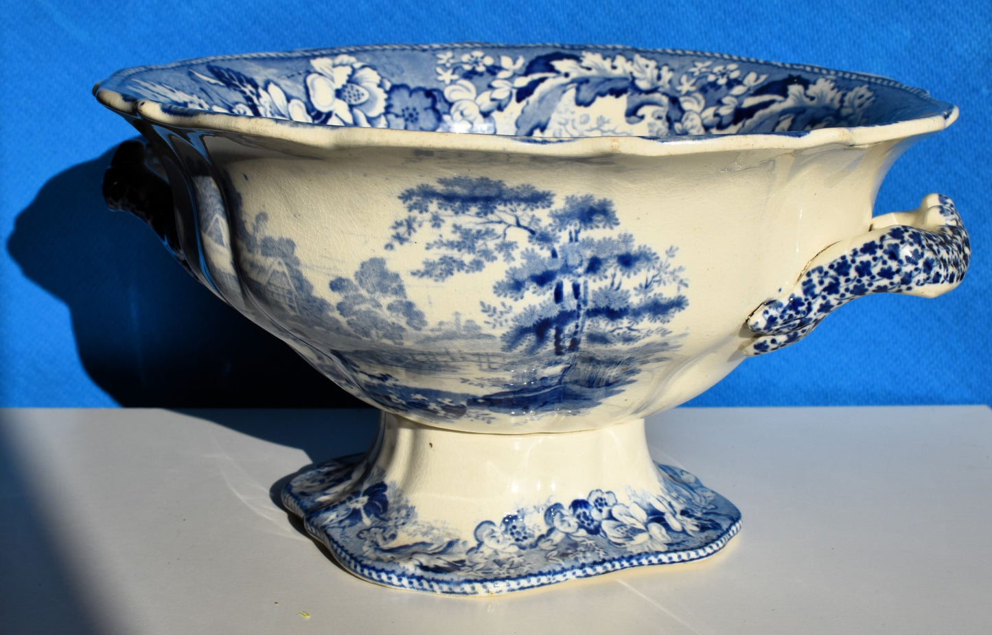 A white and blue footed bowl is decorated with country scenes and blue flowers