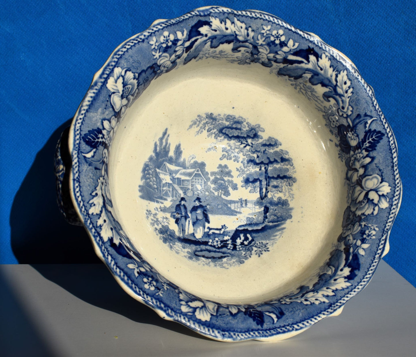 A white and blue footed bowl is decorated with country scenes and blue flowers