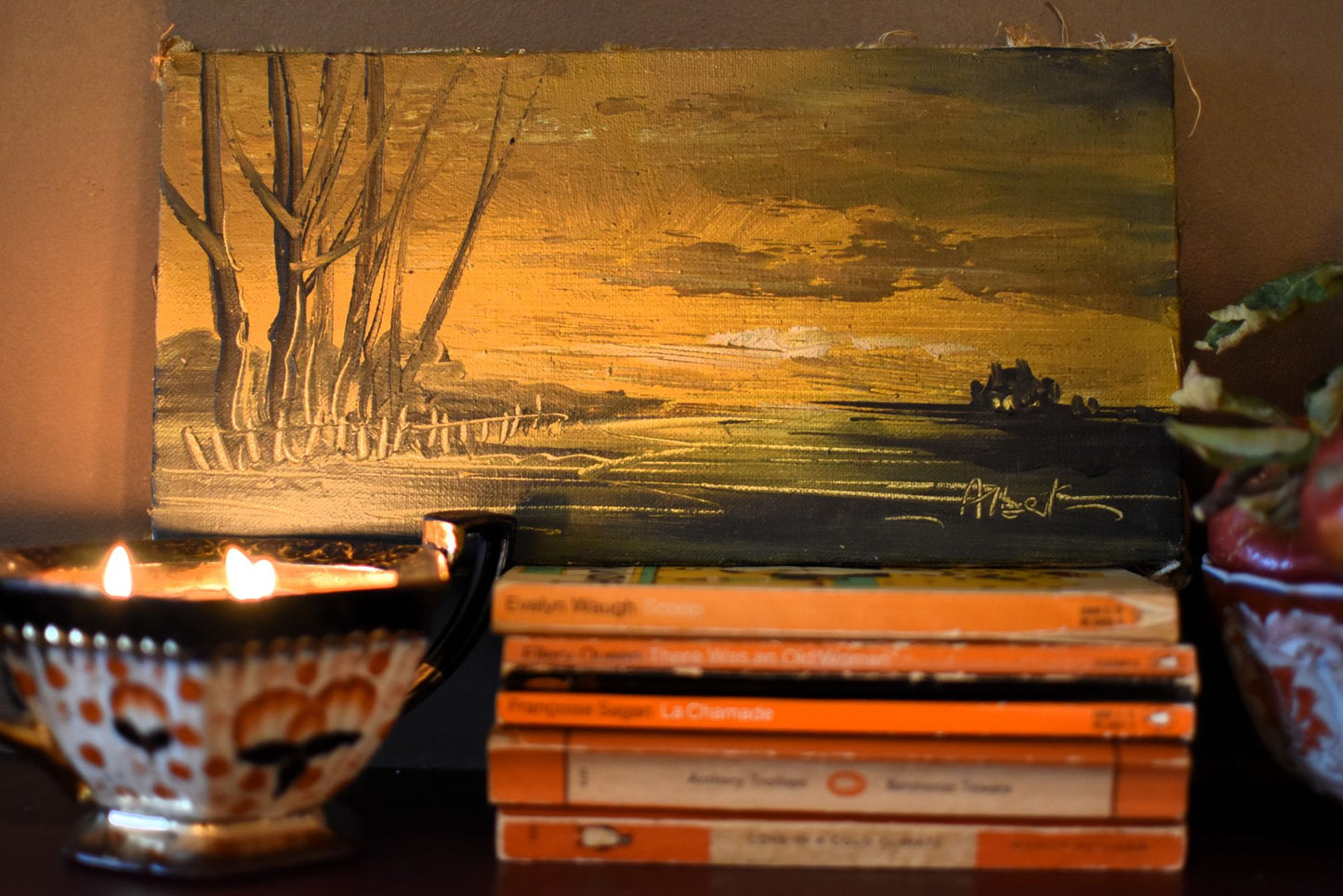 Painting of a landscape with trees and sky, placed on books with a candle beside it.