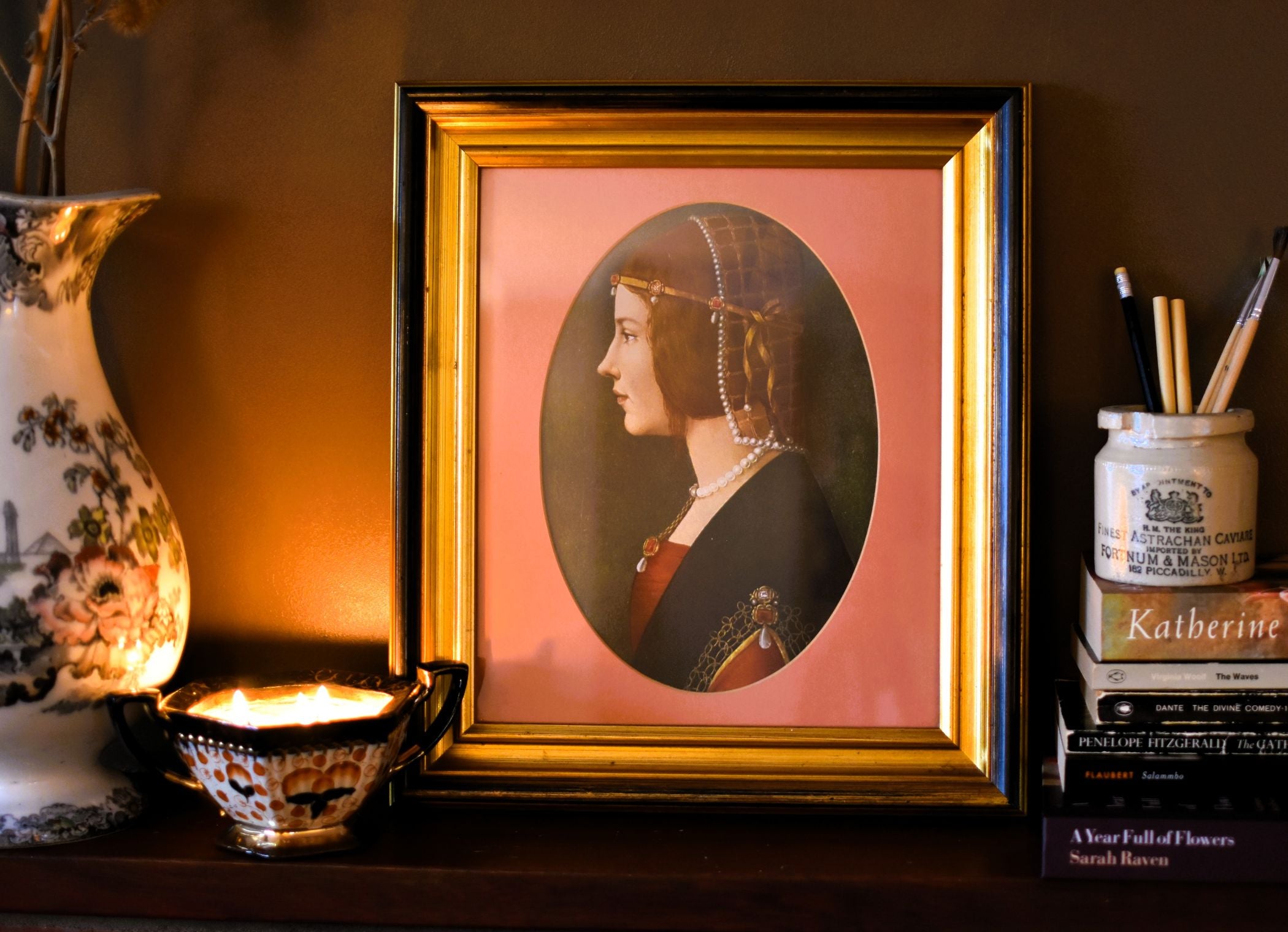 A framed print of a medieval lady wearing jewels and pearls. The frame is gold and the mount is pink