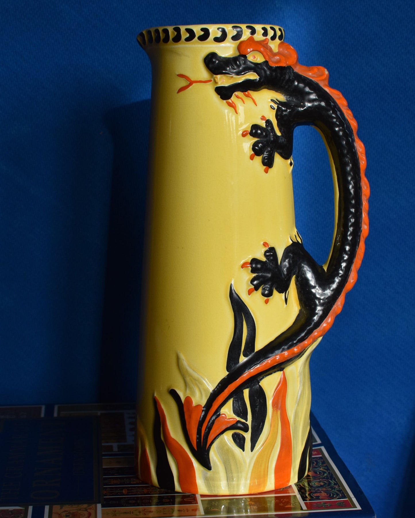 A tall yellow art deco jug decorated with a hand painted black and red dragon. The dragon's body forms the jug handle.