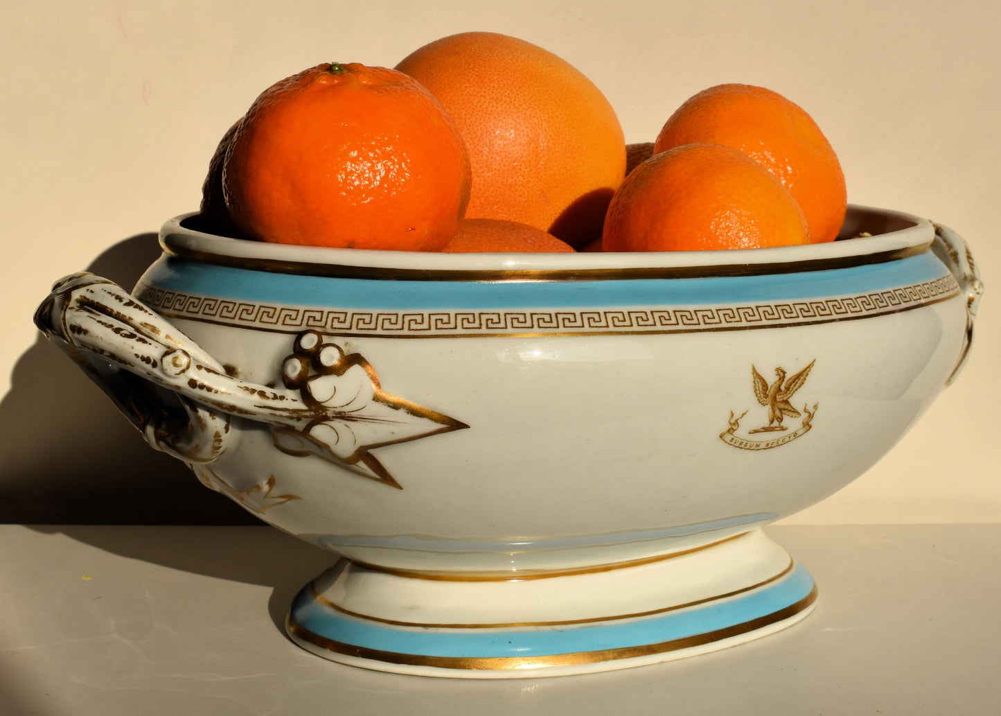 A large white tureen with gold decoration of Greek key motif and ivy leaves. The white, blue and gold bowl is full of oranges