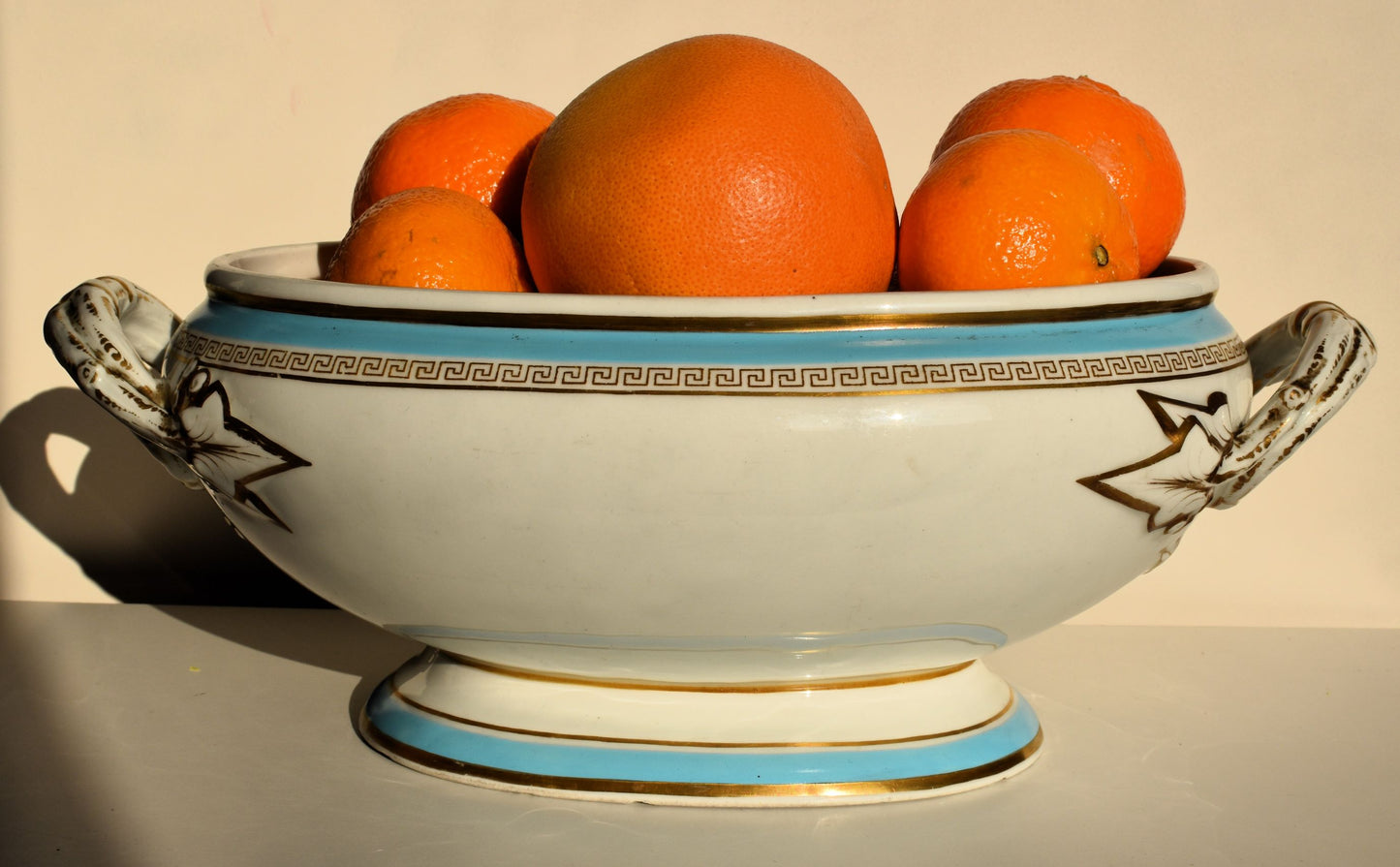 A large white tureen with gold decoration of Greek key motif and ivy leaves. The white, blue and gold bowl is full of oranges
