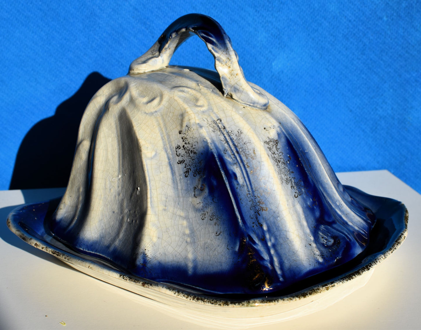 A blue and white cheese dish with a curving lid and handle is decorated with gold gilding
