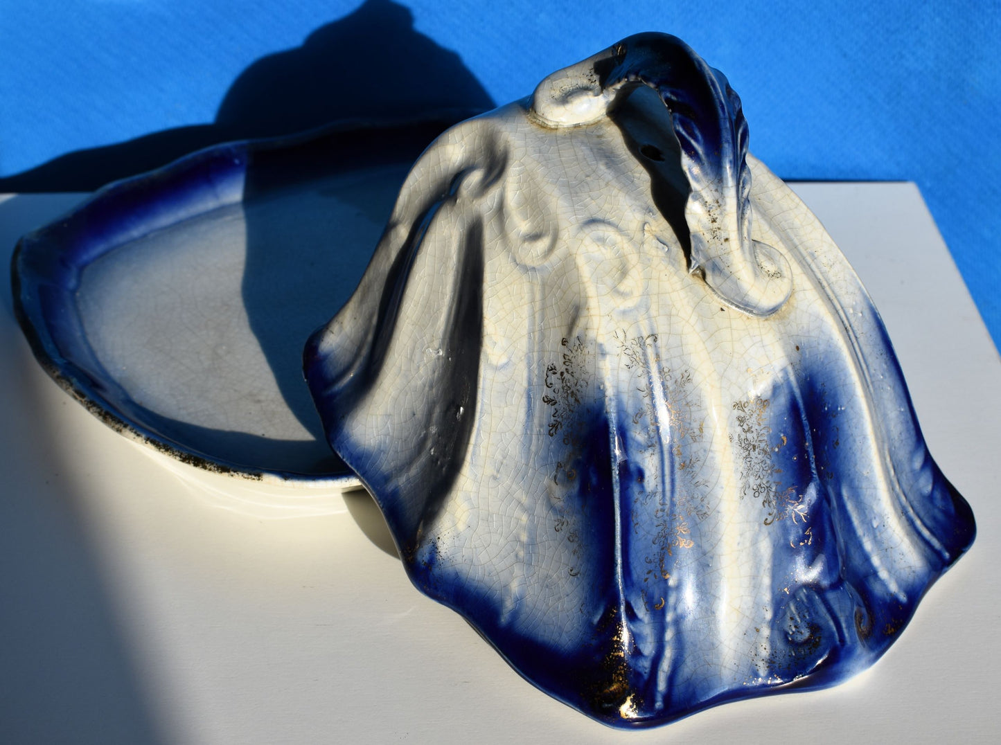 A blue and white cheese dish with a curving lid and handle is decorated with gold gilding
