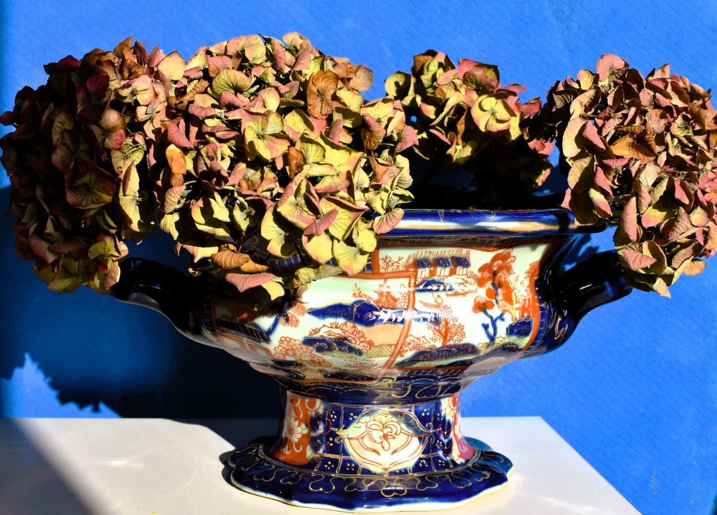 A large deep blue pedestal bowl with 2 handles is decorated in orange and gold. It holds several dried hydrangea heads