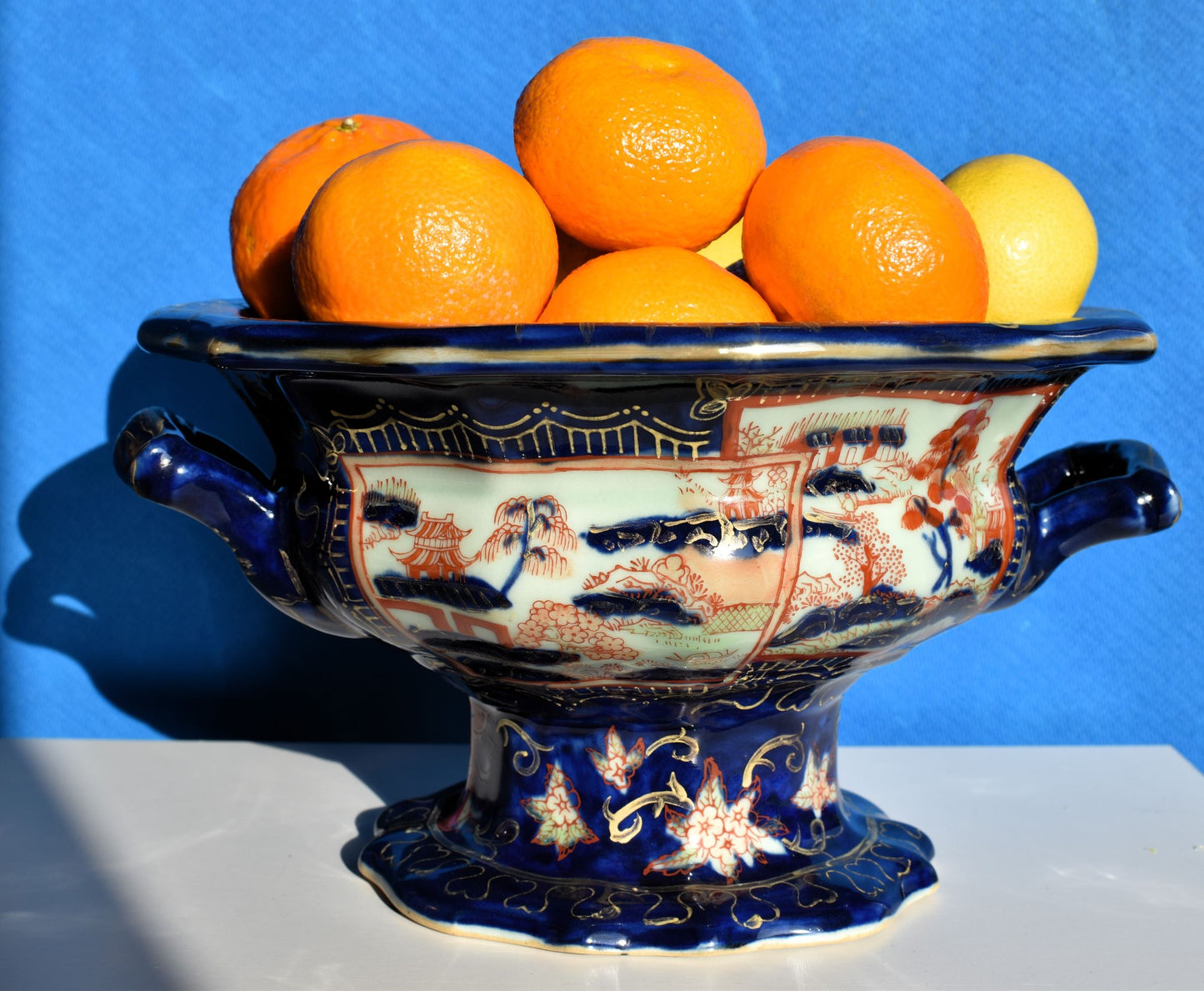 A large dark blue footed bowl has 2 handles and is decorated in orange and gold. It is full of oranges and lemons