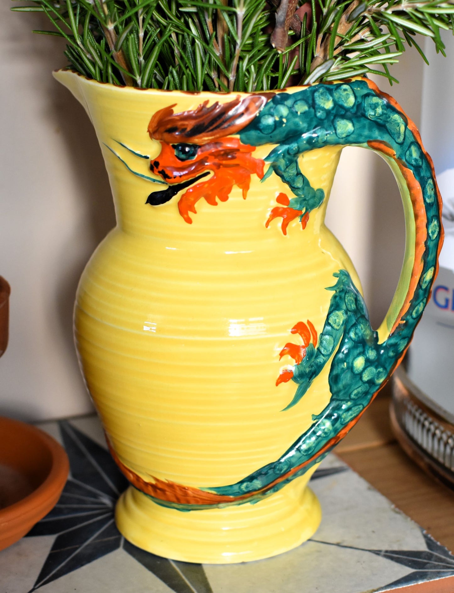 A yellow jug is decorated with an art deco dragon. The dragon's body is green and its head, tail and claws are red. The dragon's body is the handle of the jug