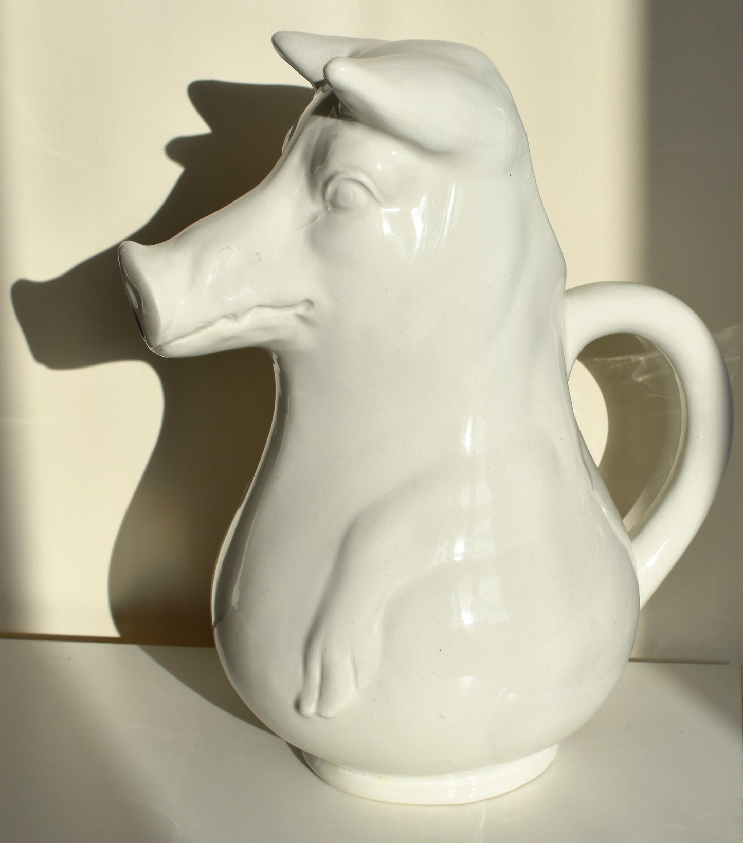 A large white jug is shaped like a pig sitting up, with its snout as the spout of the jug (other side view)
