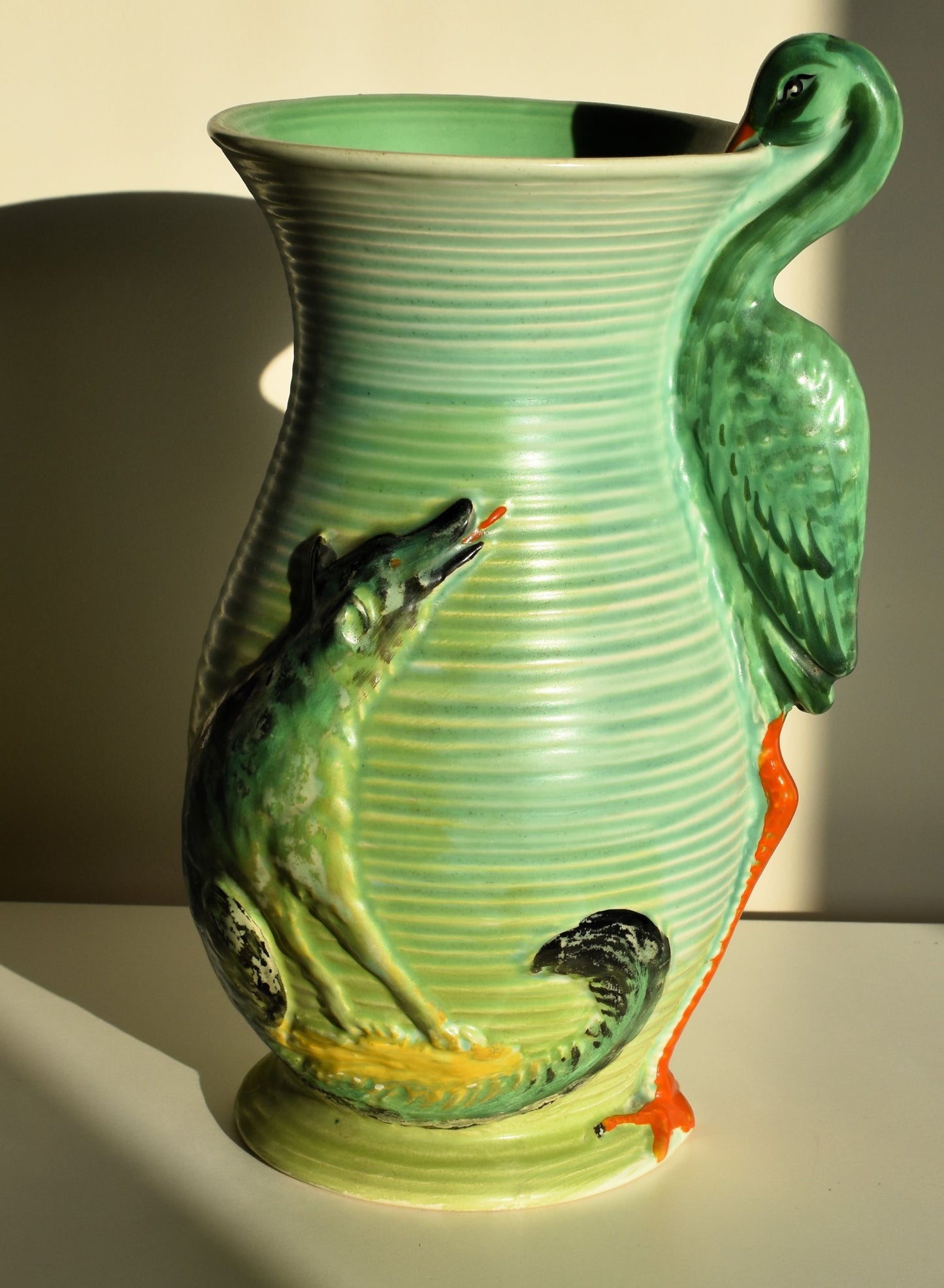 A tall green vase is decorated with a wolf watching a stork. The stork forms the handle of the vase.