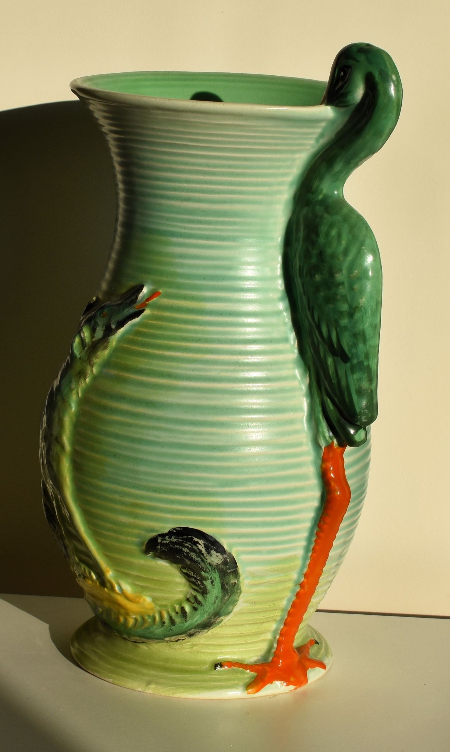 A tall green vase is decorated with a wolf watching a stork. The stork forms the handle of the vase.