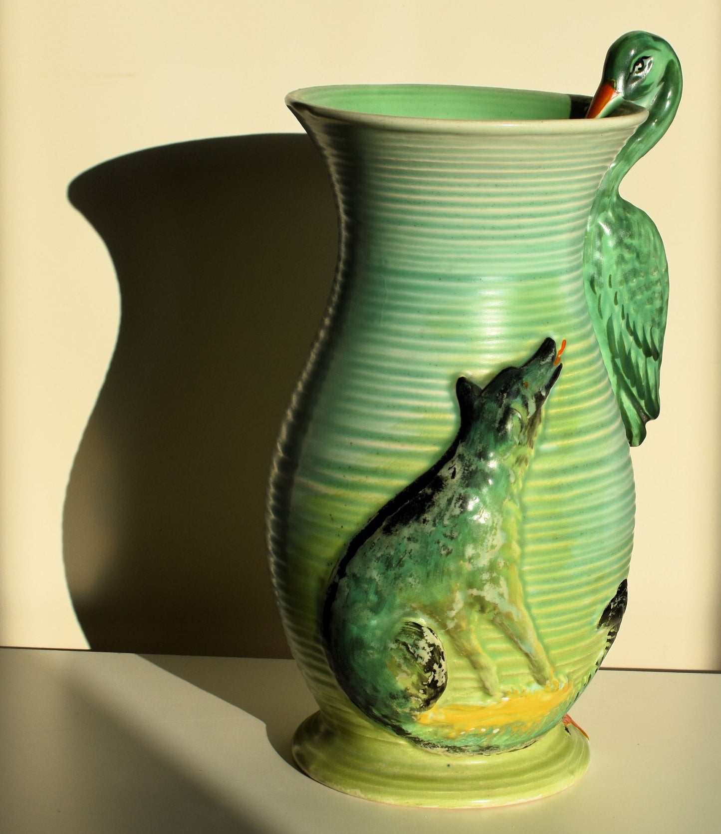 A tall green vase is decorated with a wolf watching a stork. The stork forms the handle of the vase.