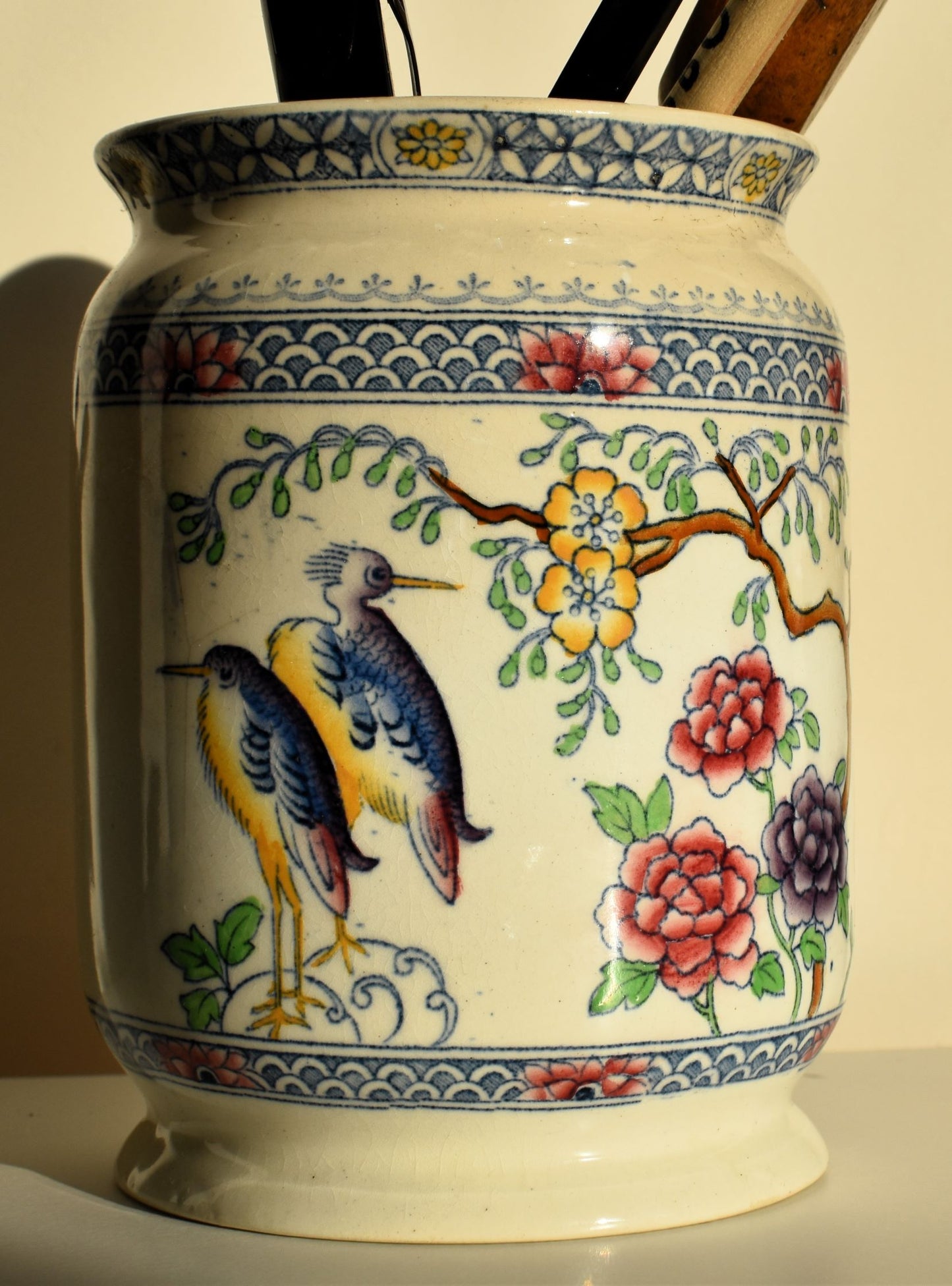A cream pot is decorated with blue and yellow storks and a branch with yellow flowers, with extra flowers in pink and purple