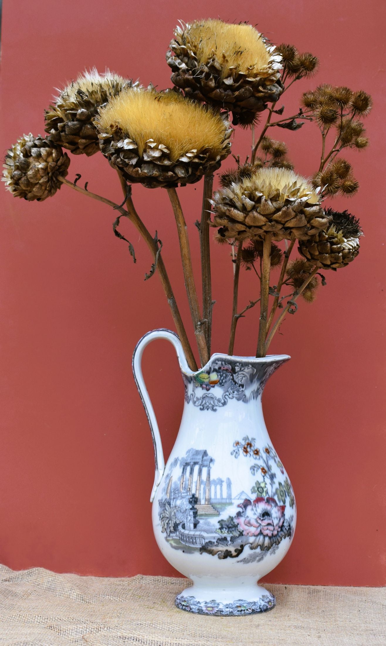 A tall white jug decorated with ancient ruins and hand painted flowers. Displays a huge dried artichoke and other seed heads