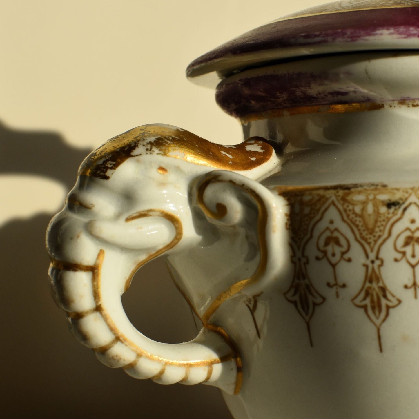 A white lidded tureen is decorated in pink and gold, it has elaborate feet and top handle and 2 side handles shaped like elephant heads and trunks