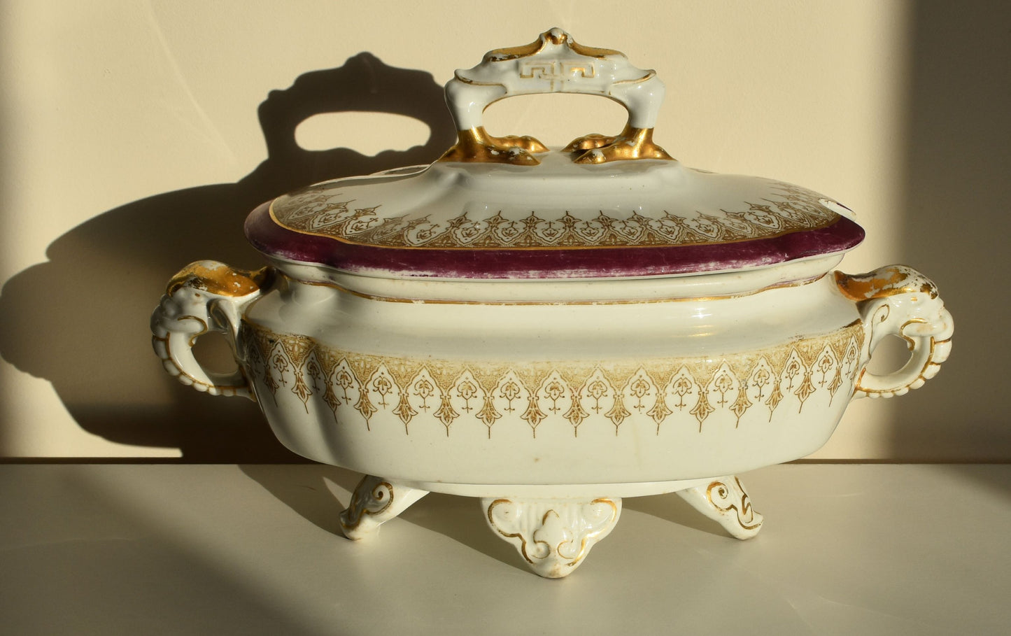 A white lidded tureen is decorated in pink and gold, it has elaborate feet and top handle and 2 side handles shaped like elephant heads and trunks