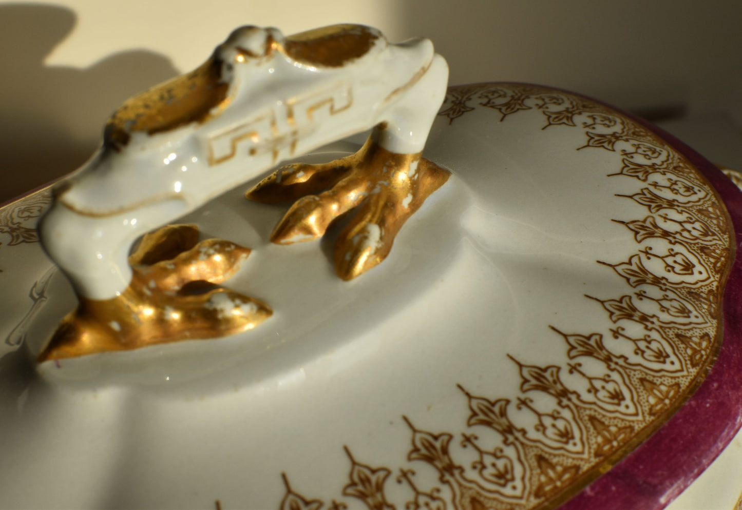 A white lidded tureen is decorated in pink and gold, it has elaborate feet and top handle and 2 side handles shaped like elephant heads and trunks