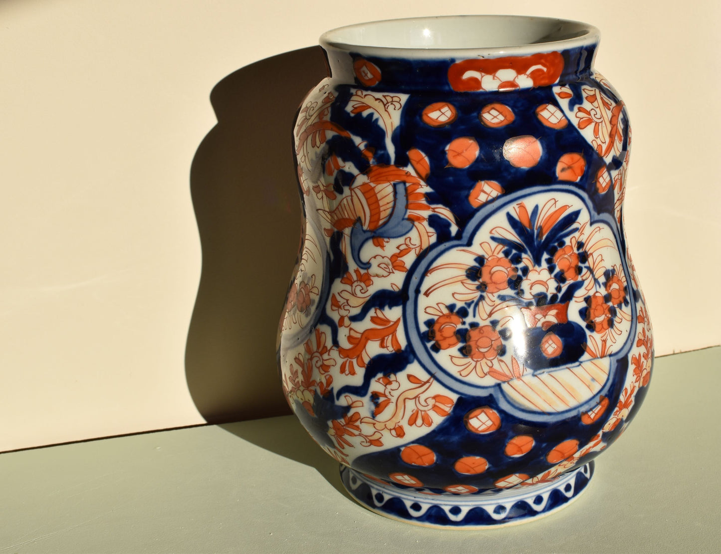 A large, squat vase in the imari colours of orange and blue, with a central floral arrangement and other geometric patterns painted around the sides. the Inside of the vase is white