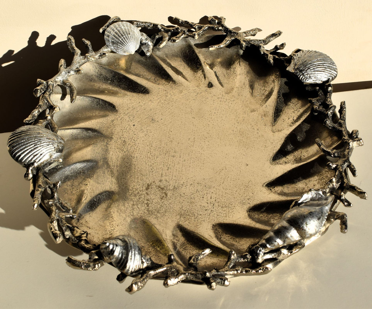 A large round metal dish is decorated with silver metal clamshells, spiral shells and twisted coral