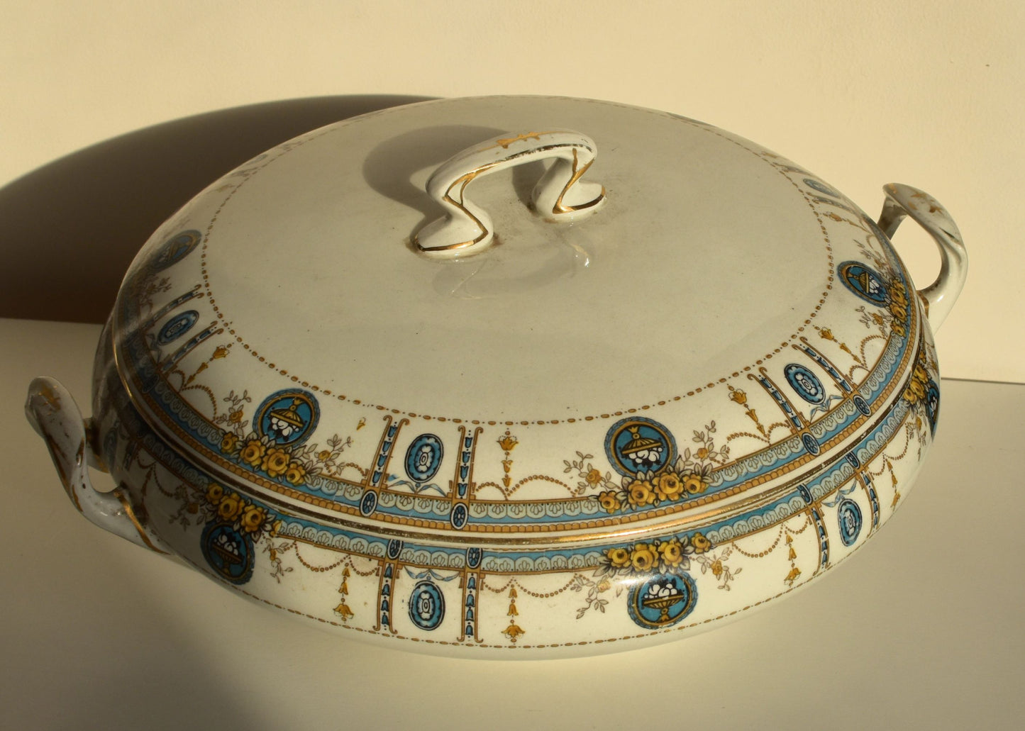 A wide, shallow white tureen is decorated with green and gold swags and flowers over the lid and sides. It has 2 handles