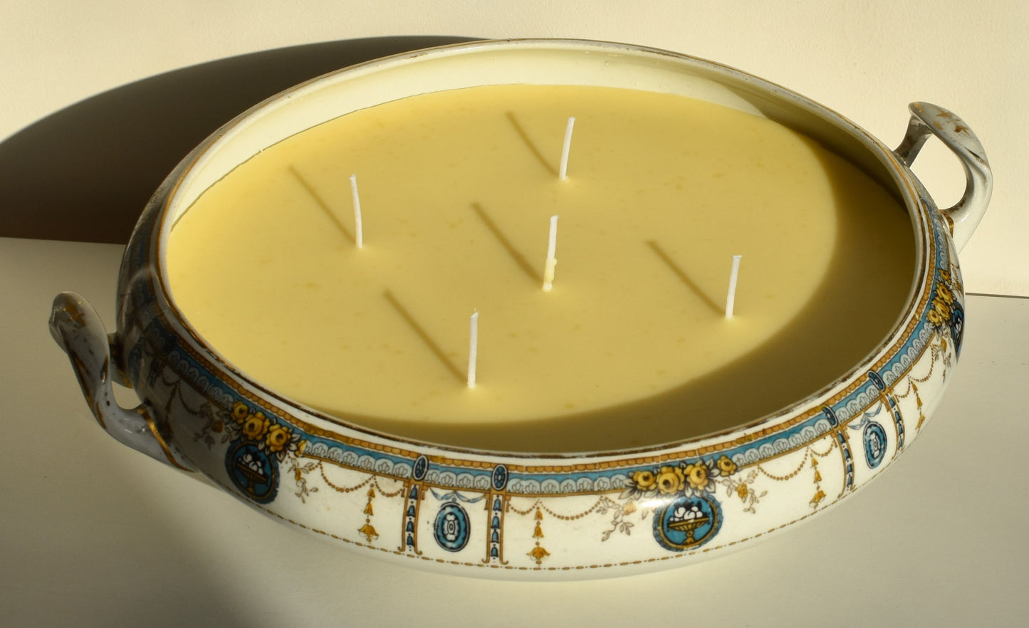 A wide, shallow white tureen is decorated with green and gold swags and flowers. It's filled with golden wax and 5 wicks to form a big candle