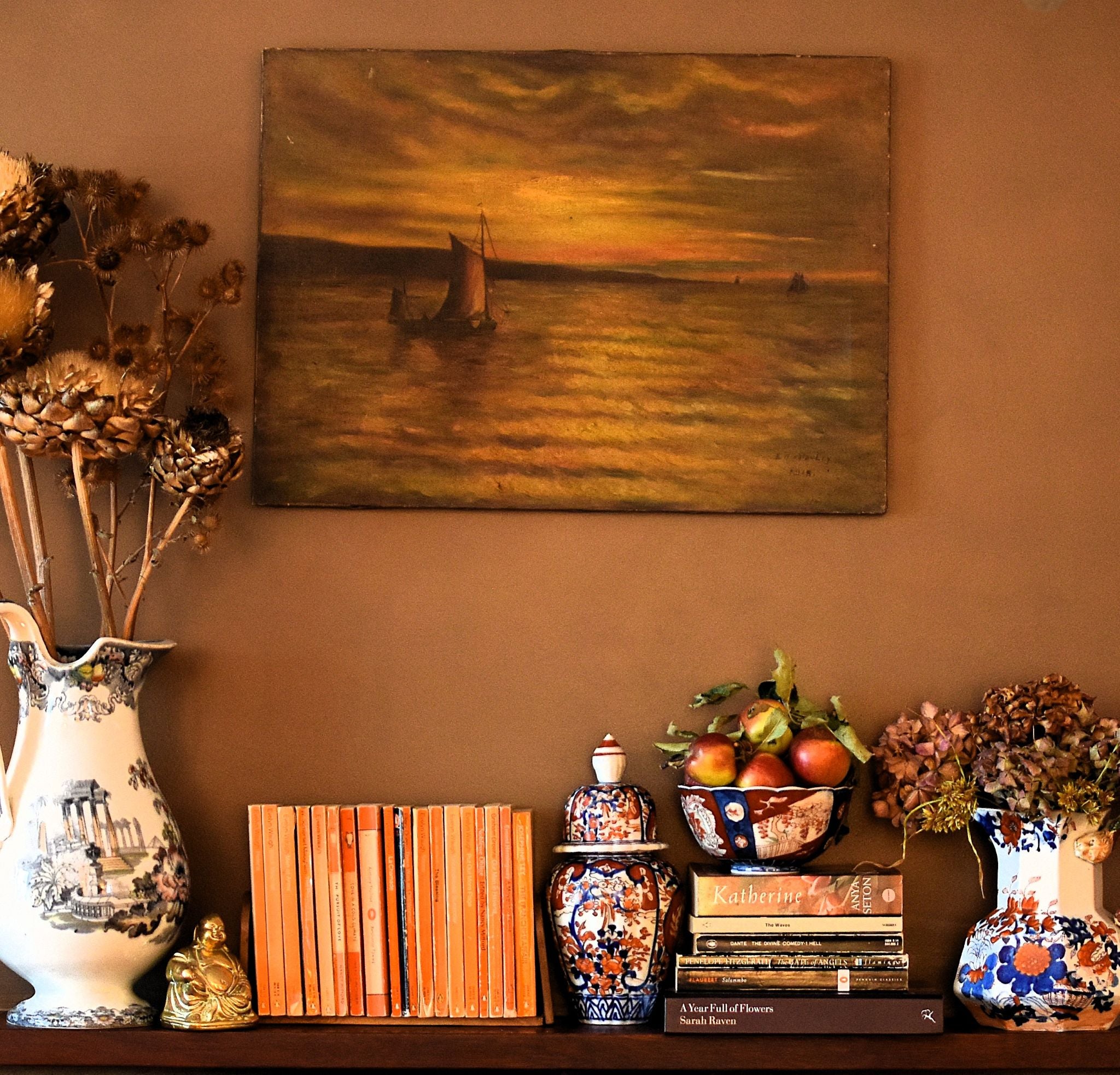Decorative shelf with books, vases, and a painting of a sunset over water on a brown wall.