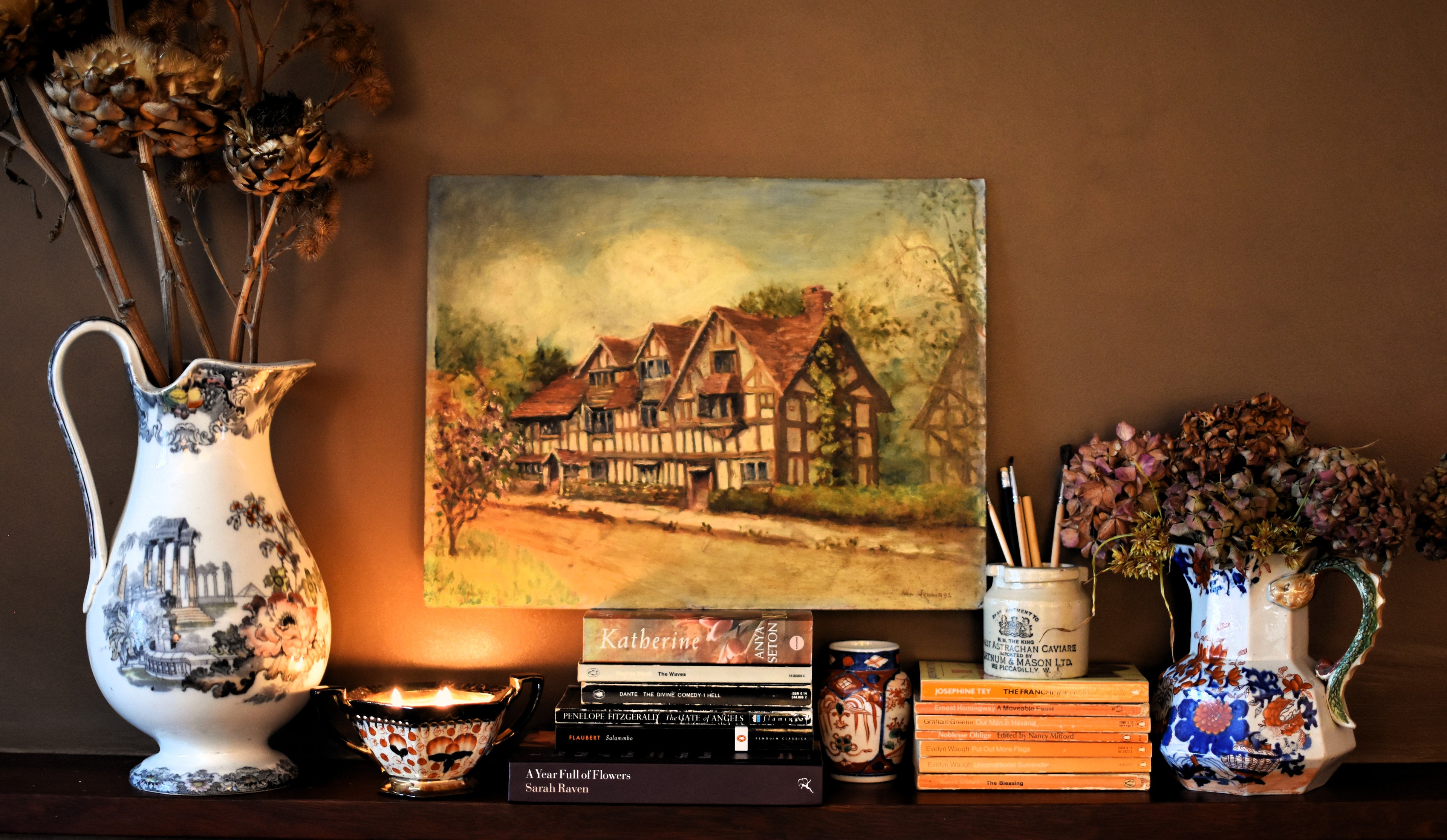 Decorative shelf with a painting of a house, vases, and books against a brown wall.