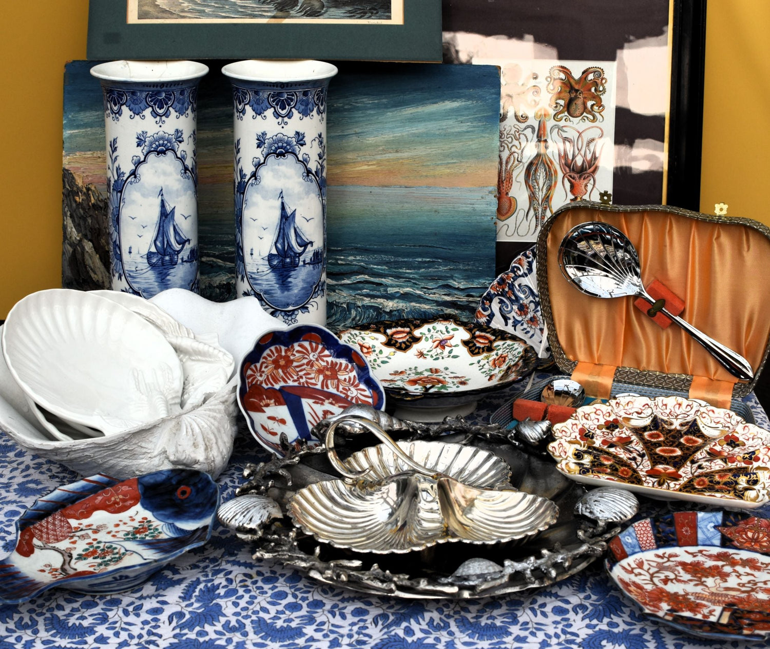 shell shaped plates and silver dishes and spoons are arranged in front of blue and white vases and oil paintings of the sea