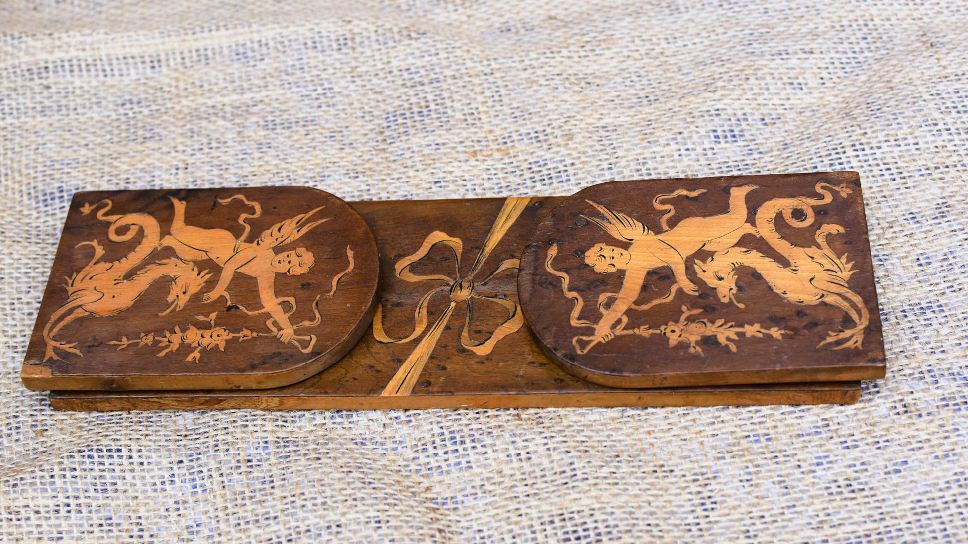Wooden decorative panel with intricate carvings of cherubs and ribbons on a textured fabric background.