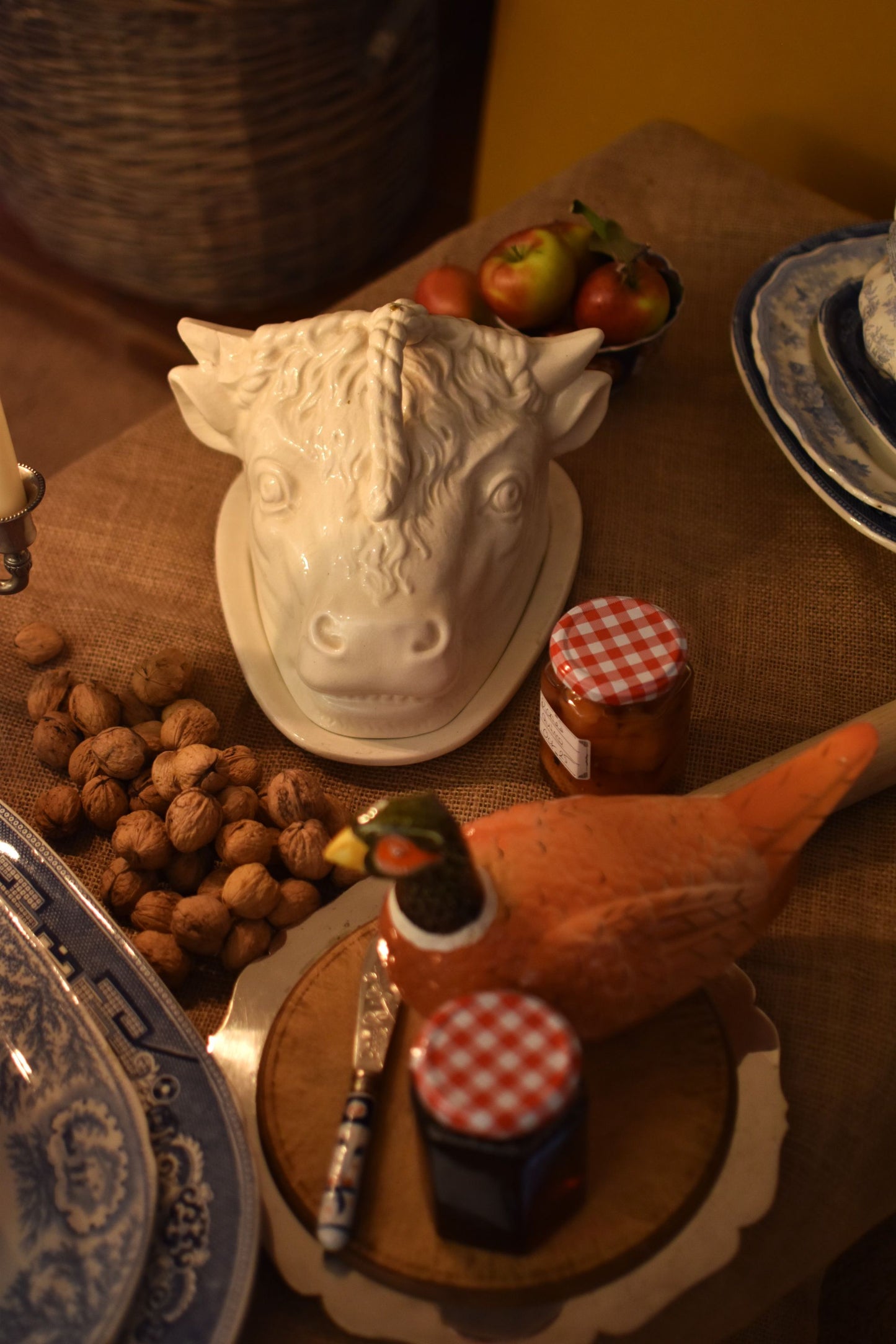 A serving dish in the shape of a bull or cow's head in the middle of a table also showing jam, walnuts and apples on a brown hessian 