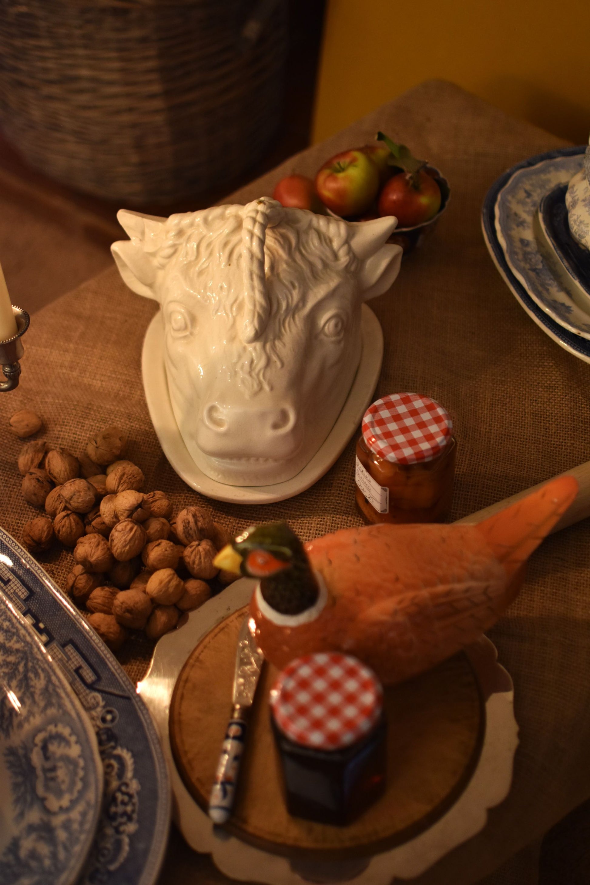 A serving dish in the shape of a bull or cow's head in the middle of a table also showing jam, walnuts and apples on a brown hessian 