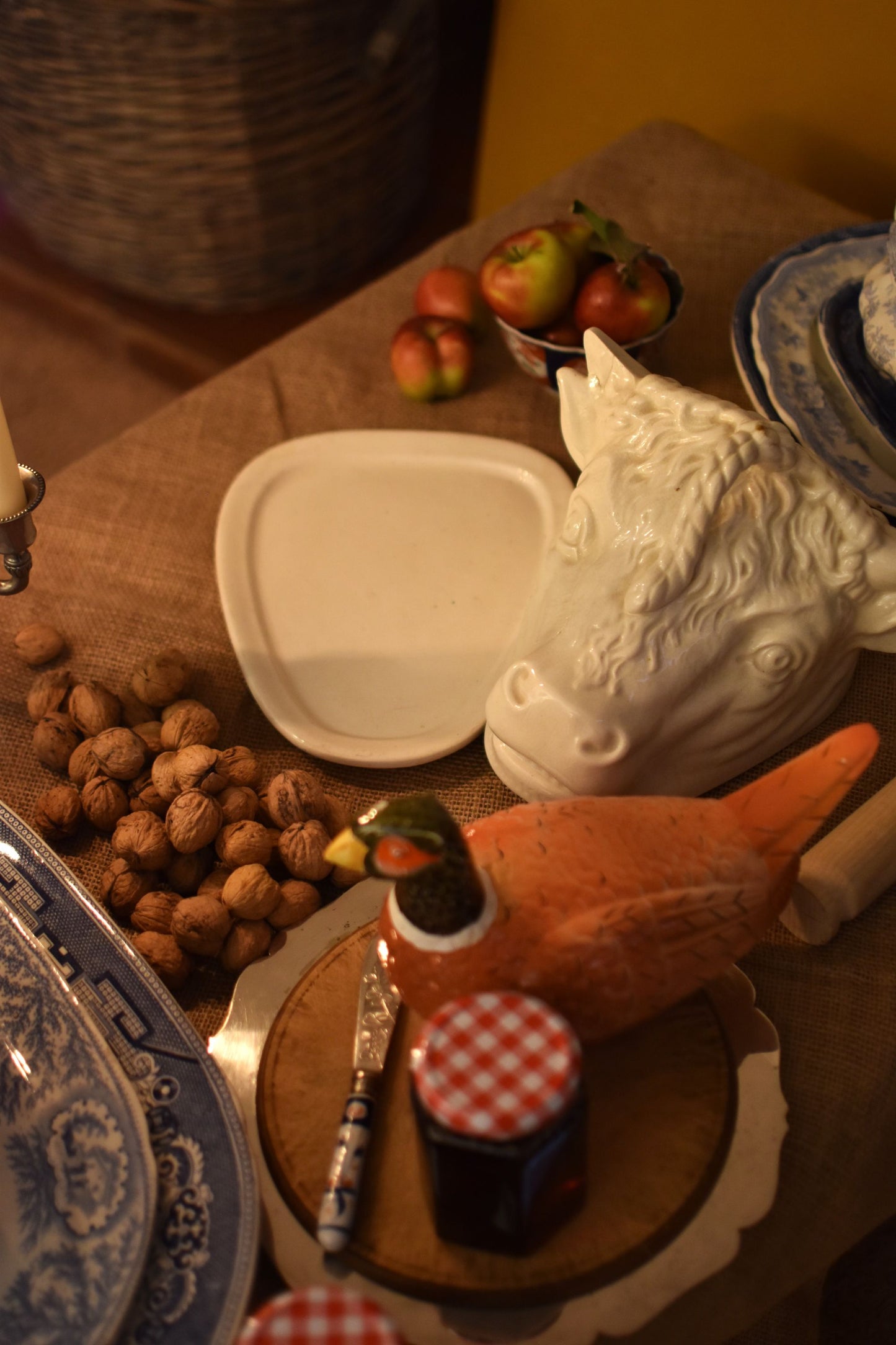 A serving dish in the shape of a bull or cow's head in the middle of a table also showing jam, walnuts and apples on a brown hessian 