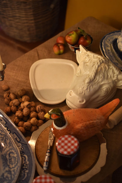 A serving dish in the shape of a bull or cow's head in the middle of a table also showing jam, walnuts and apples on a brown hessian 