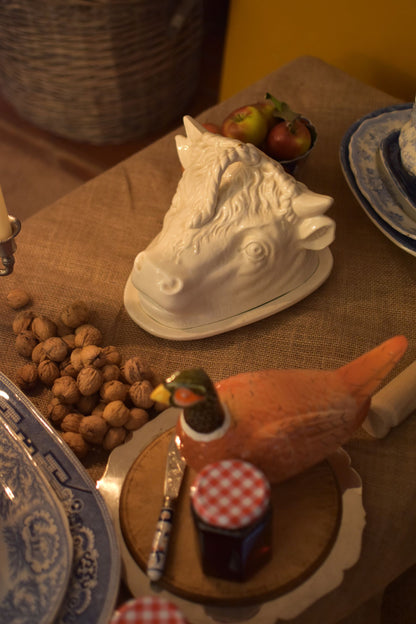 A serving dish in the shape of a bull or cow's head in the middle of a table also showing jam, walnuts and apples on a brown hessian 