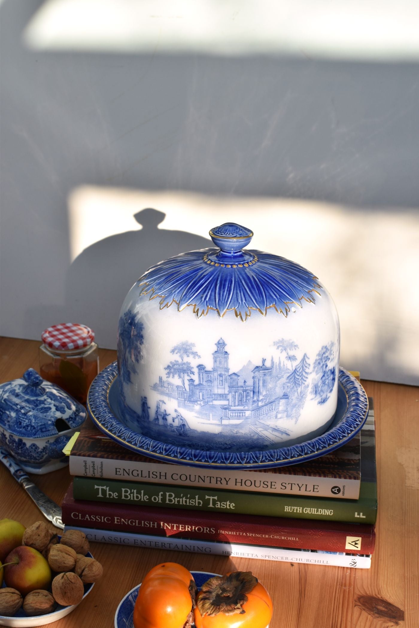 Blue and white ceramic cloche on a stack of books with fruit and a jar on a wooden surface.