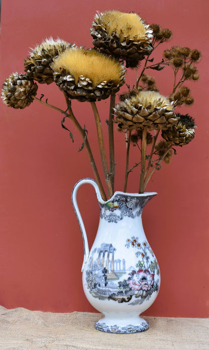 A tall white jug decorated with ancient ruins and hand painted flowers. Displays a huge dried artichoke and other seed heads