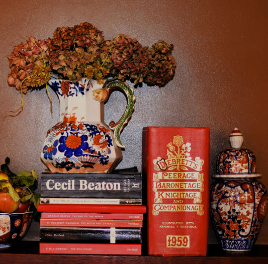A large white and imari floral jug sits on top of a pile of books, next to a big red book called Debretts peerage