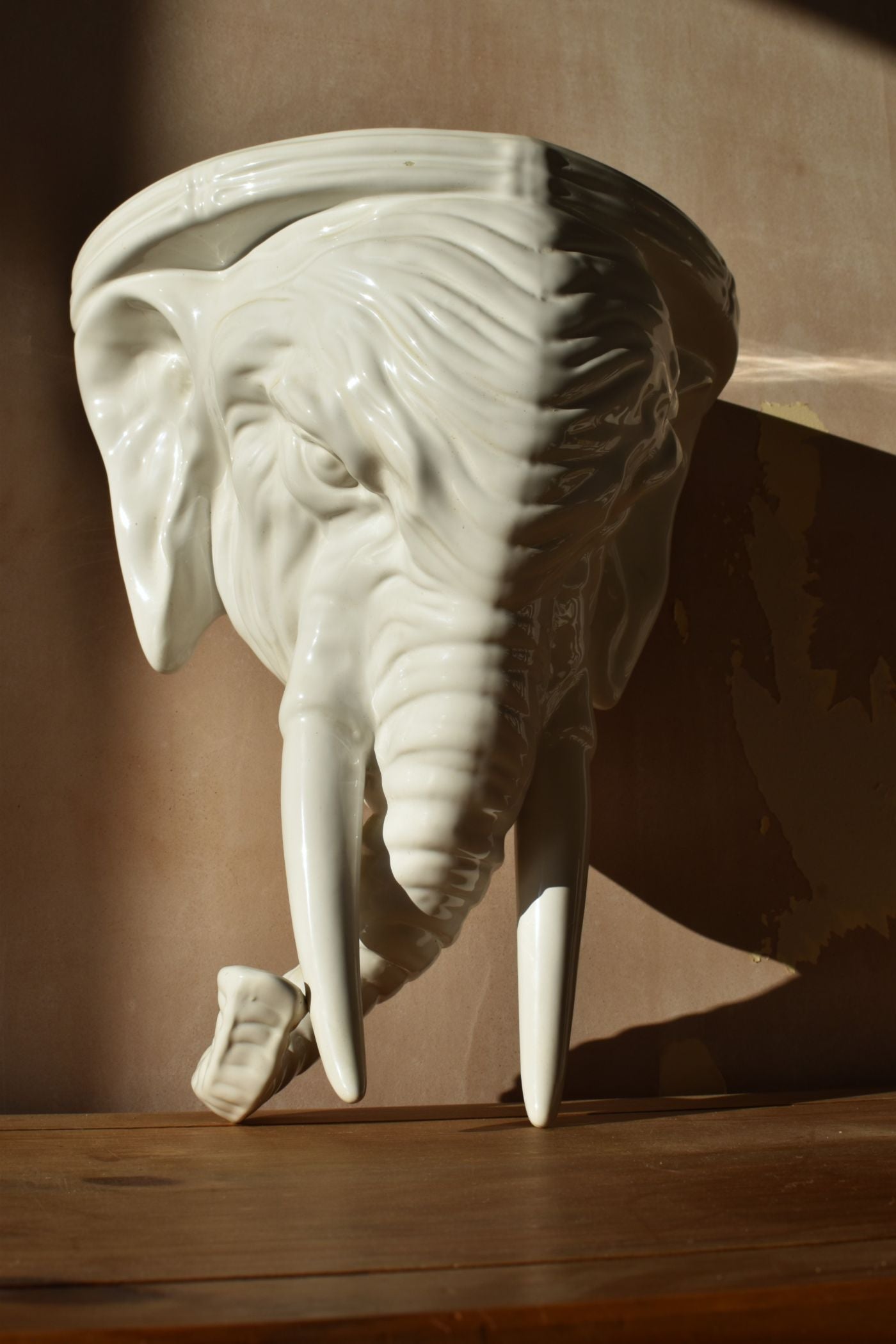 A large white elephant made of porcelain, with two long tusks and a curled trunk, is against a pink background and half in shadow
