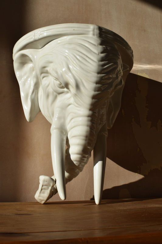 A large white elephant made of porcelain, with two long tusks and a curled trunk, is against a pink background and half in shadow