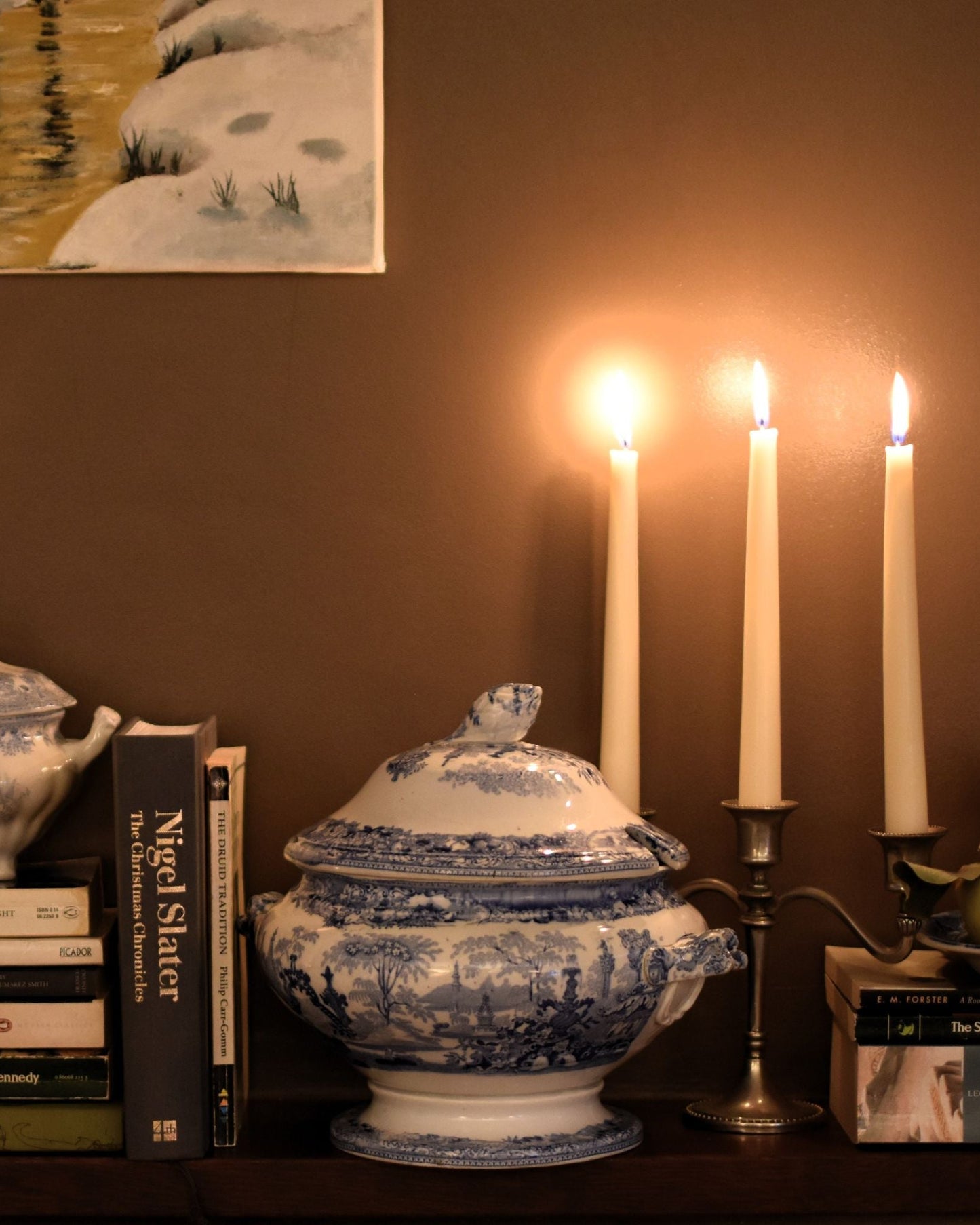 A blue and white bowl with handles and lid is on a shelf with books and candles, against a brown wall
