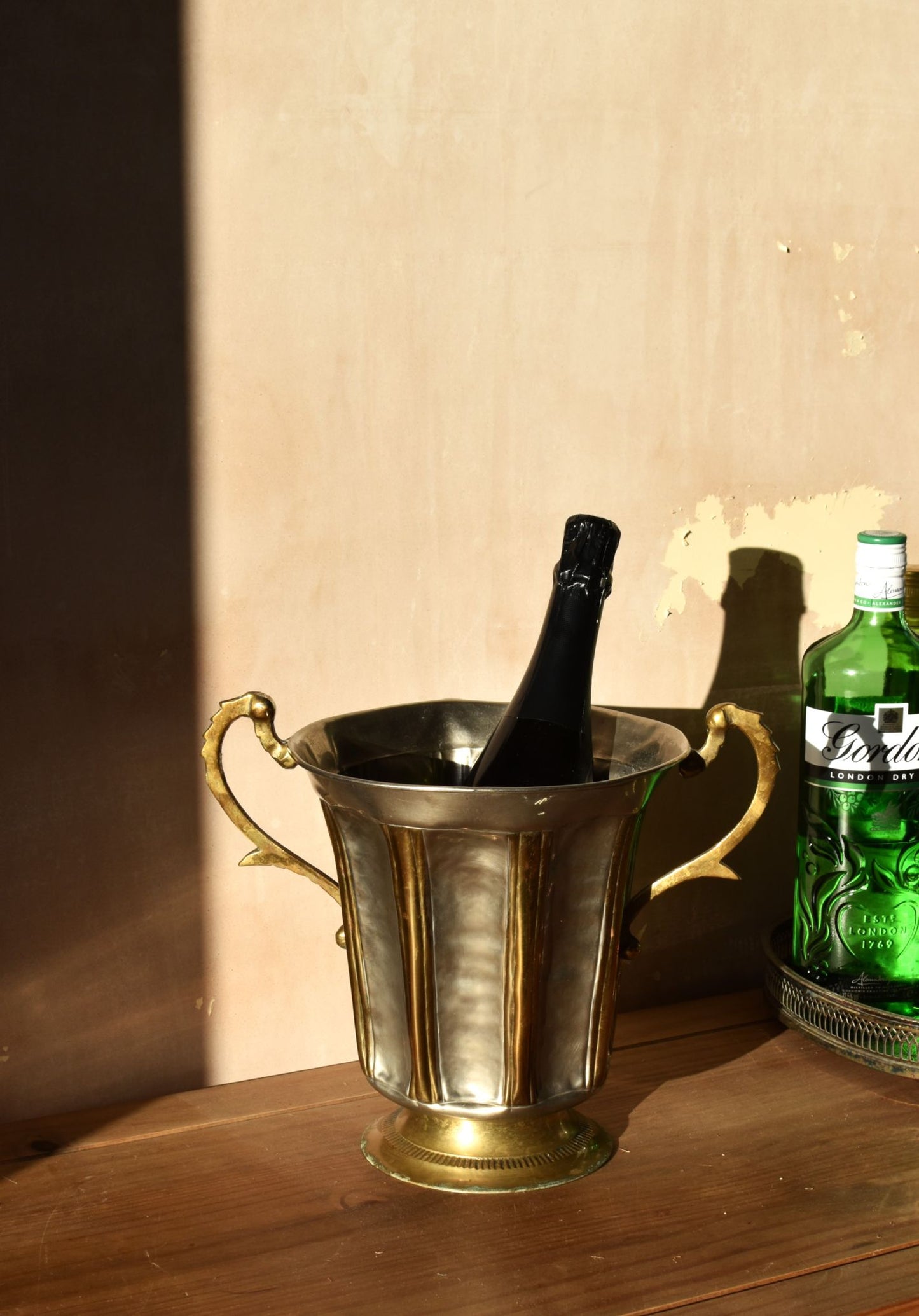 a silver and gold ice bucket with big handles is on a wooden shelf. it has a bottle of champagne inside