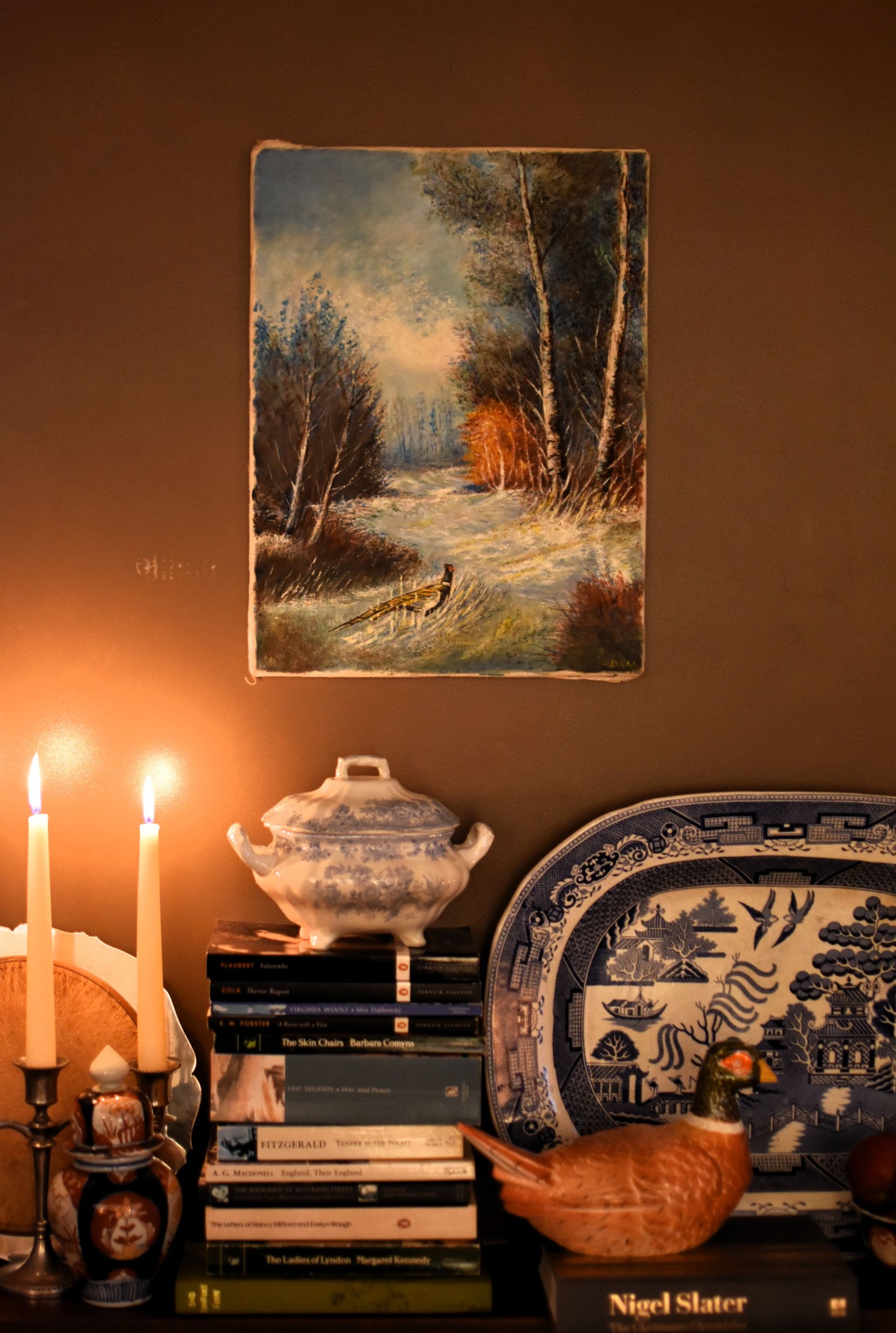 Decorative shelf with books, candles, and a painting of a forest scene on a brown wall.