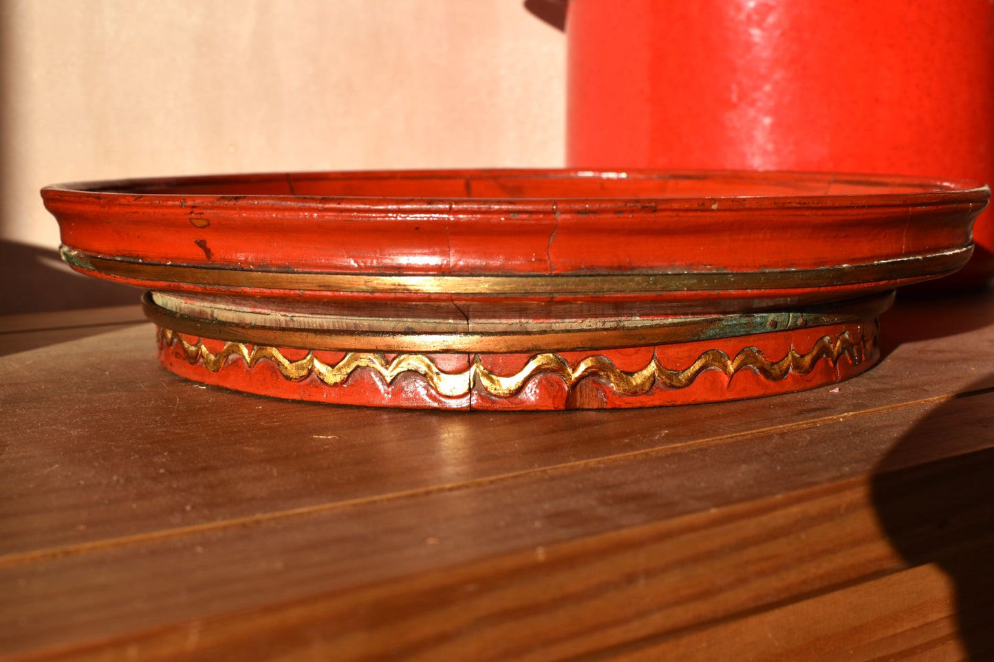 red painted wooden raised plate has bands of gold coloured metal around the edge and base, on a wood table