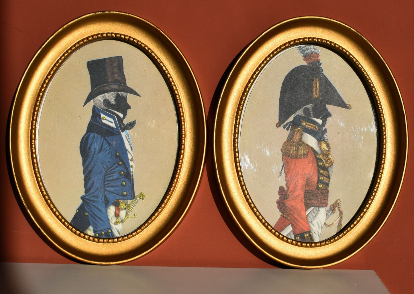Two silhouette paintings in gold oval frames. The silhouettes each show a man in military uniform from about 1803, the left is blue and the right is red