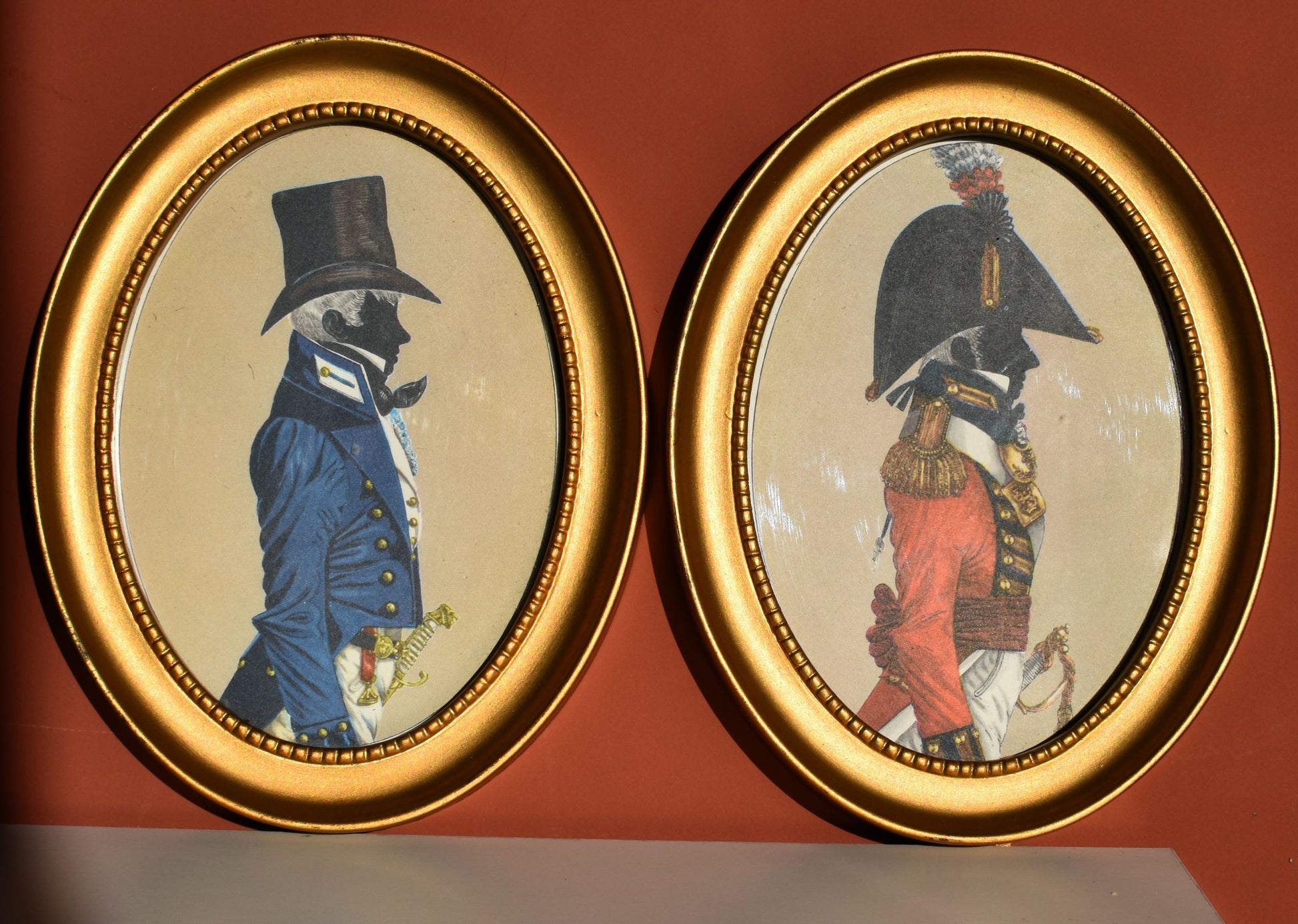 Two silhouette paintings in gold oval frames. The silhouettes each show a man in military uniform from about 1803, the left is blue and the right is red