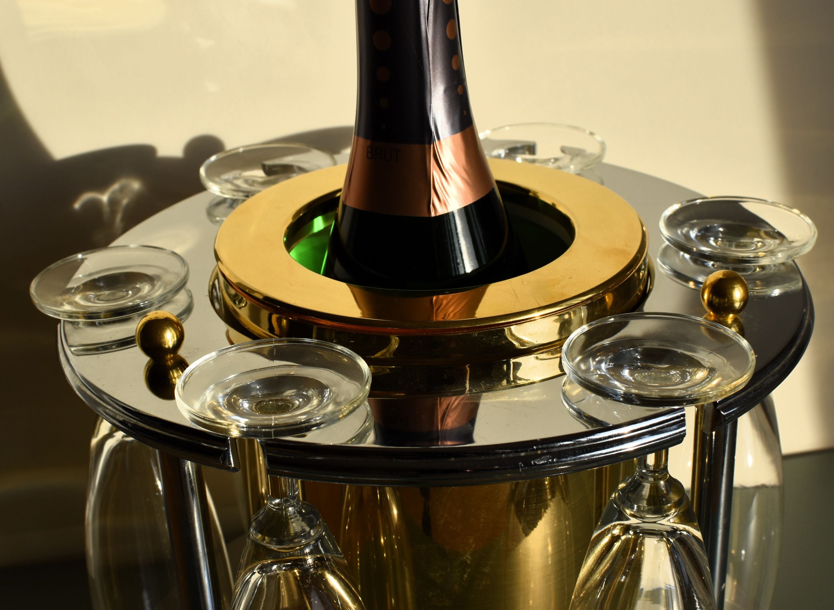 a brass and chrome ice bucket with a bottle of champagne in, with 6 tall champagne glasses hanging from it against a white background