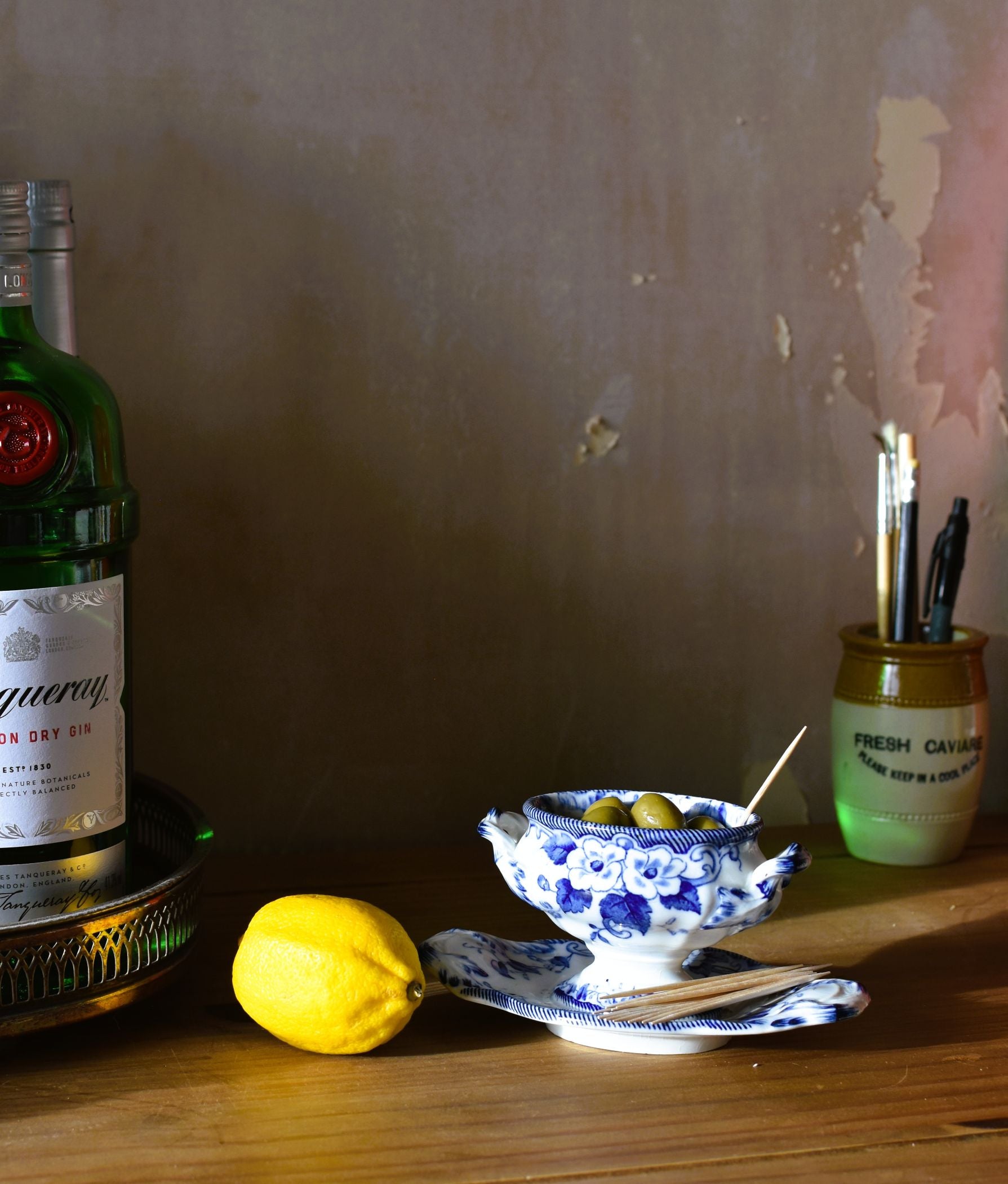 a blue and white dish full of olives is on a wooden table with a lemon and bottles of drinks