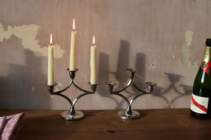 a silver candelabra on a wooden table with three white candles, next to an idential silver candelabra with no candles