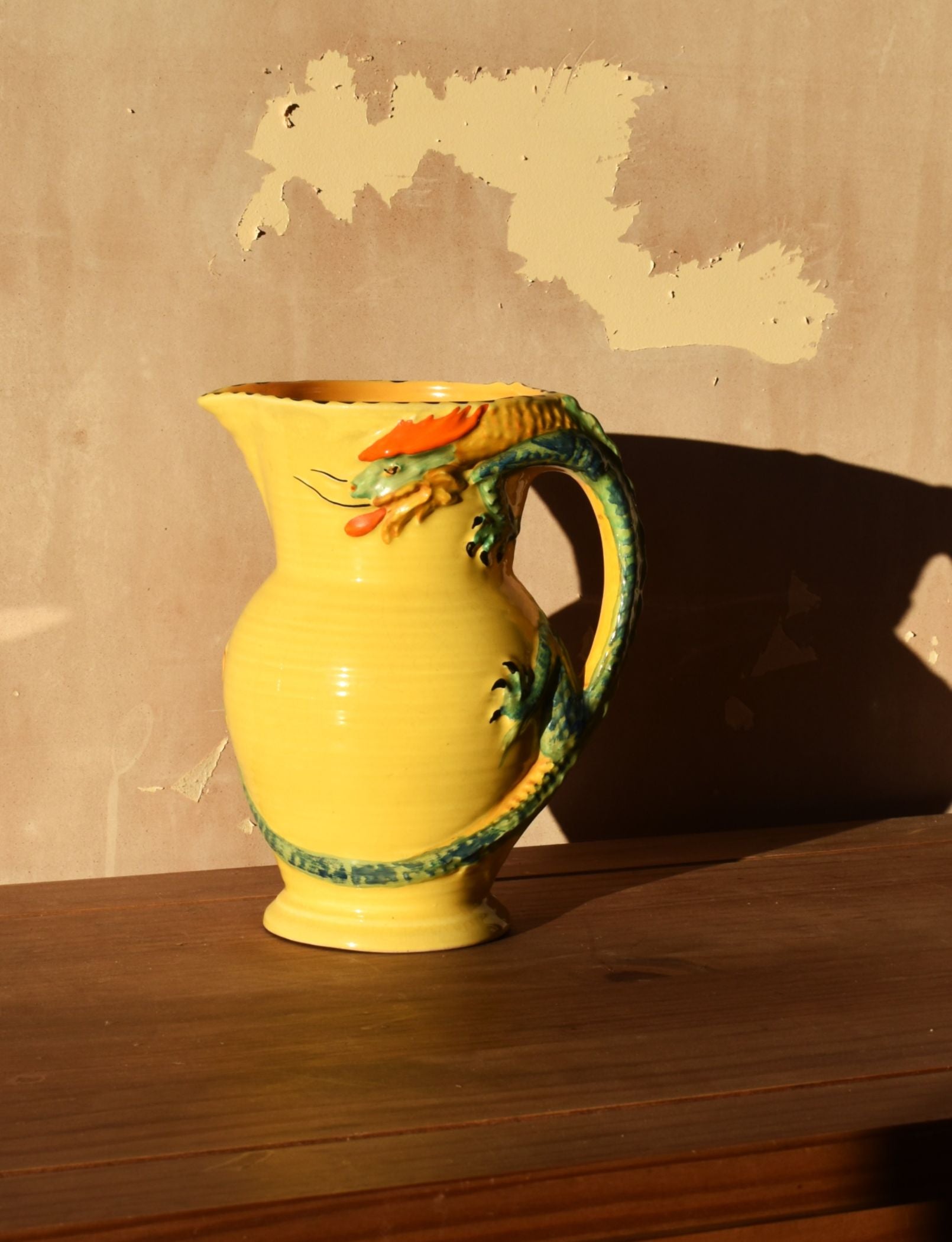 Yellow pitcher with dragon design on a wooden surface against a beige wall.