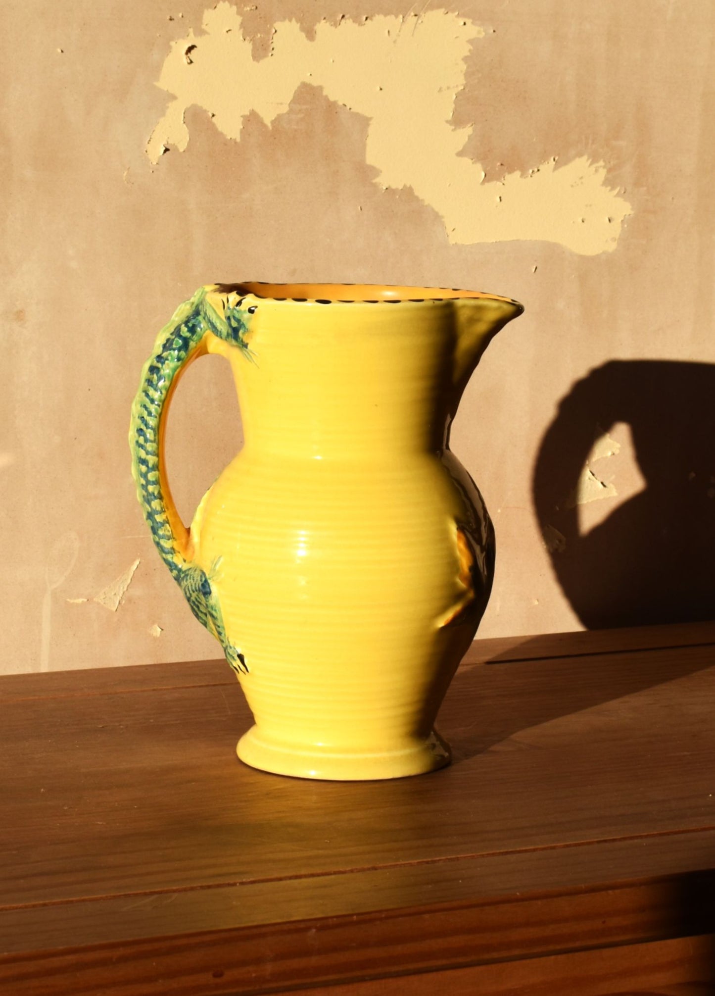 Yellow pitcher with a green handle on a wooden surface against a beige wall.